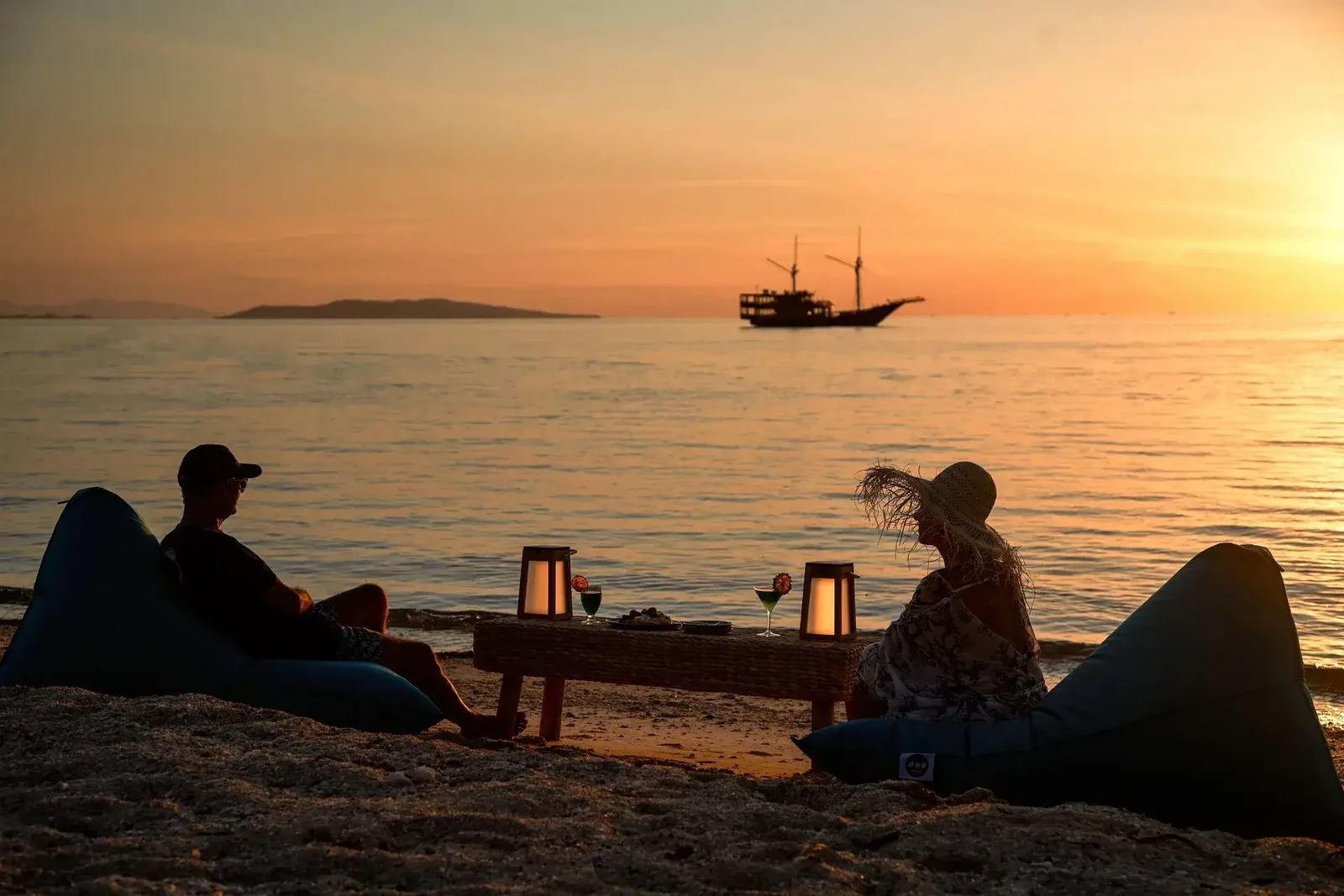 Guests at a beach sundowner setup with the liveaboard anchored offshore at sunset in Komodo